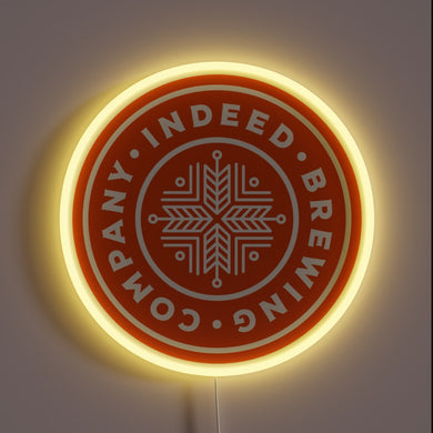 indeed-brewing-company-yellowpop-93558 – Round neon sign with an orange background and white snowflake-style logo for Indeed Brewing Company, outlined in soft yellow glow