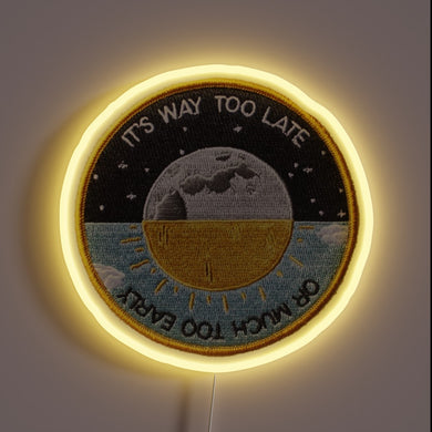 Alt text: Circular neon patch sign with moon and sun illustration, reading “It’s Way Too Late or Much Too Early.”

