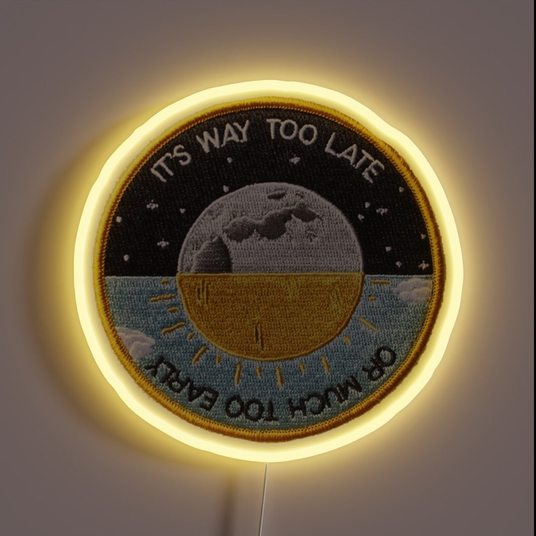 Alt text: Circular neon patch sign with moon and sun illustration, reading “It’s Way Too Late or Much Too Early.”
